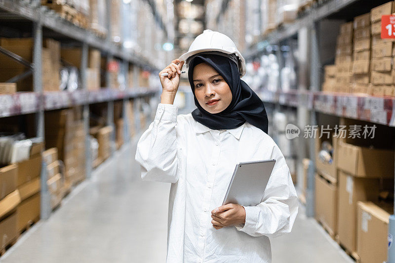 Portrait of Asian muslim female headscarf hijab wear hard hat holding digital tablet checklist product shelves warehouse. woman worker inspecting goods industry. Logistic concept.